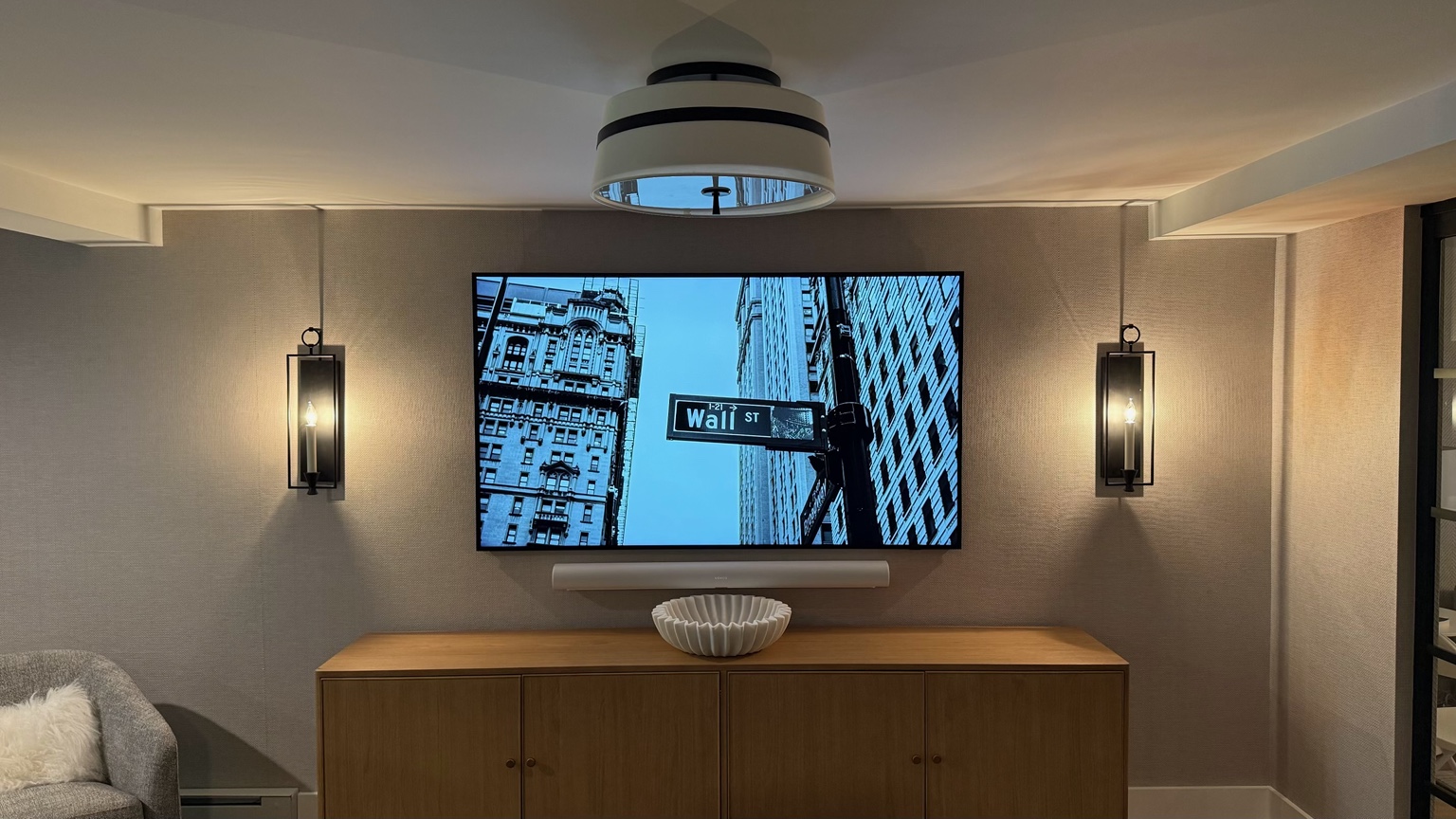 Wall-mounted TV and soundbar above a wood cabinet, framed by wall sconces.